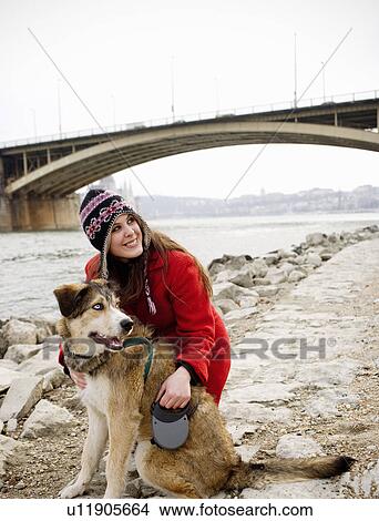 Picture - Young woman kneeling by dog on riverside, smiling. Fotosearch
