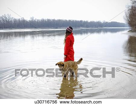 Stock Photograph - Young woman standing in river with dog surrounded by ripples. Fotosearch