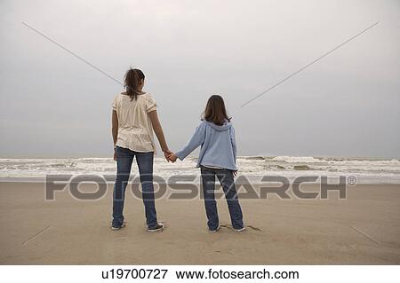 Stock Photo - Mother and daughter (9-11) holding hands on beach, rear view. Fotosearch