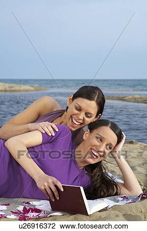 Stock Image - Two young women on beach one is reading to the other smiling sea and sky in background.. Fotosearch