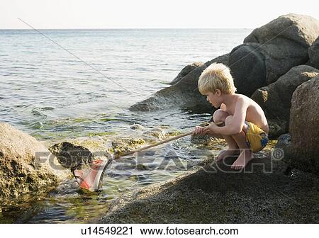 Stock Image - Young boy crouched on rocks fishing with a net.. Fotosearch