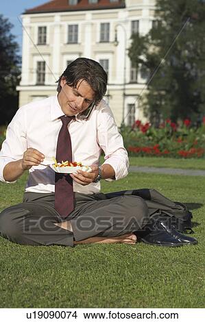Picture - Businessman on square sitting in meadow on the mobile whilst eating chips laughing.. Fotosearch
