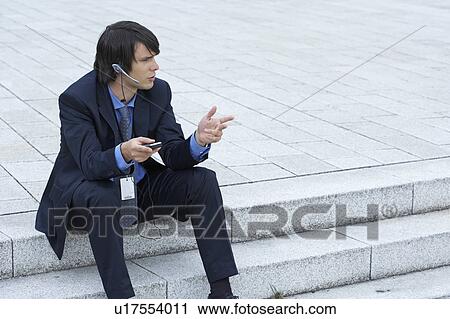 Stock Image - Businessman sitting on steps with headset and mobile discussing looking into distance.. Fotosearch