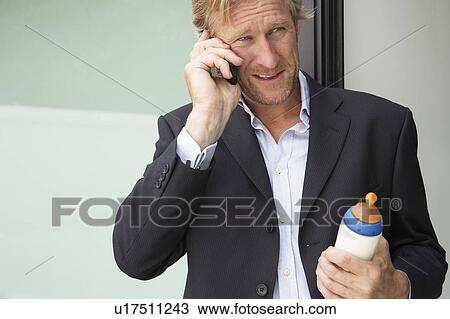 Stock Image - Businessman using mobile phone whilst holding baby's bottle.. Fotosearch