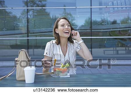 Stock Photograph - Businesswoman on table in front of office outside on the mobile whilst eating salad to go looking up smiling.. Fotosearch