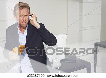 Stock Photo - Man in office eating croissant on telephone with reflections.. Fotosearch