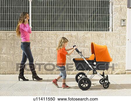 Young girl (5-7) pushing buggy on pavement while mother talks on mobile phone, Alicante, Spain, View Large Photo Image Stock Image - Young girl (5-7) pushing buggy on pavement while mother talks on mobile phone, Alicante, Spain,. Fotosearch