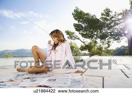 Stock Image - Woman relaxing by infinity pool reading newspaper and drinking coffee.. Fotosearch