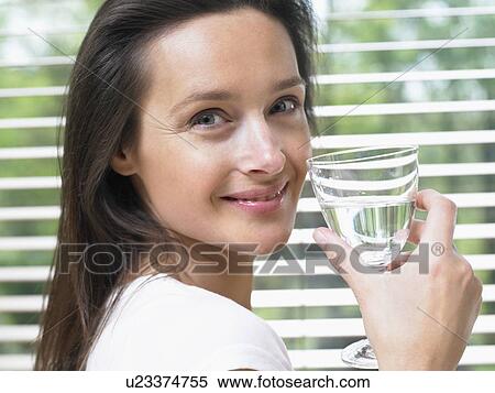Stock Photography - Woman standing by a window holding a glass of water smiling.. Fotosearch