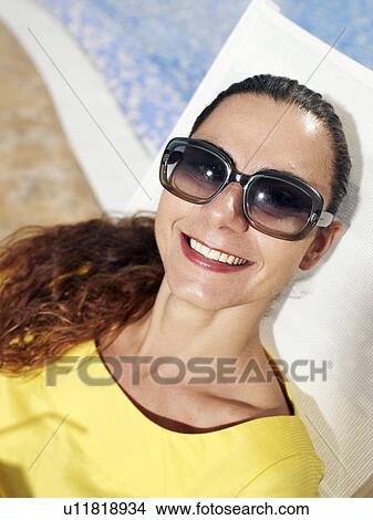Woman wearing sunglasses relaxing on sun lounger, smiling View Large Photo Image Picture - Woman wearing sunglasses relaxing on sun lounger, smiling. Fotosearch
