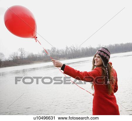 Stock Image - Young woman standing by river holding red balloon, smiling. Fotosearch