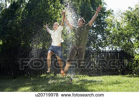 Stock Photo - Man and women jumping over garden sprinkler.. Fotosearch