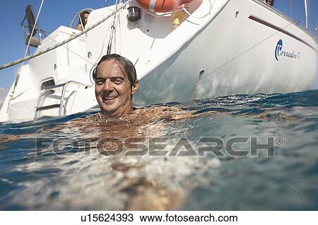 Stock Image - Mature man swimming in sea by yacht, smiling, portrait. Fotosearch
