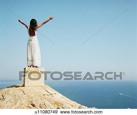 Stock Photo - Woman standing on a large cement block with arms outstretched looking out over the sea.. Fotosearch