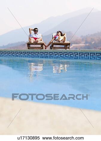 Stock Photography - Couple sunbathing by swimming pool, mountains in distance. Fotosearch