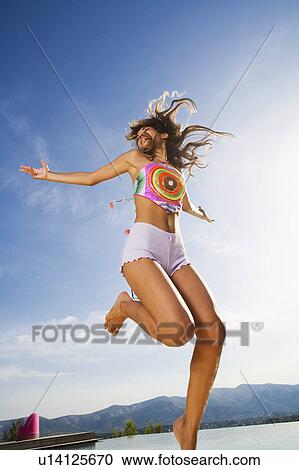 Stock Image - Woman jumps in air arms out wearing shorts outside.. Fotosearch