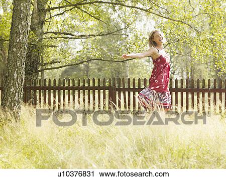 Stock Image - Woman running in grass by a wooden fence smiling.. Fotosearch