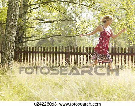 Stock Image - Woman running in grass by a wooden fence smiling.. Fotosearch