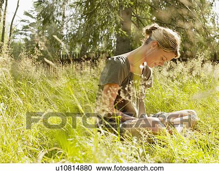 Stock Image - Woman sitting in the grass reading a book smiling.. Fotosearch