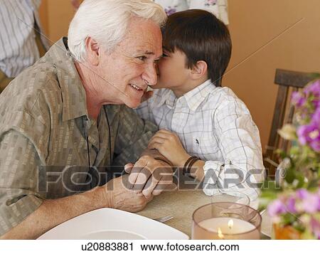 Stock Image - Boy (8-10) whispering in grandfather's ear at dining table. Fotosearch