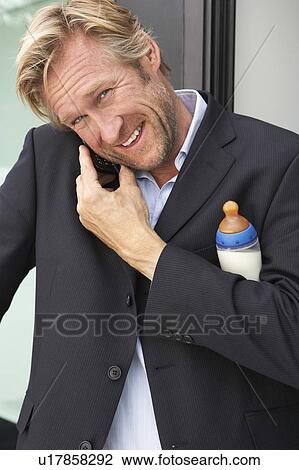 Stock Image - Businessman using mobile phone whilst holding baby's bottle.. Fotosearch