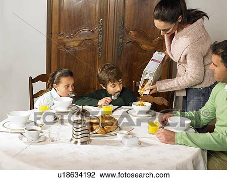 Stock Image - Family having breakfast, mother pouring cereal, boy (6-8) and girl (4-6) watching. Fotosearch