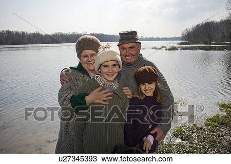 Stock Image - Grandparents standing by river with grandson (12-14) and grangdaughter (11-13) smiling. Fotosearch