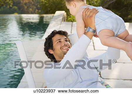Stock Photo - Man holding up a baby by infinity pool smiling.. Fotosearch