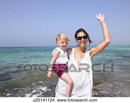 Picture - Woman holding a young girl at the beach waving and smiling.. Fotosearch