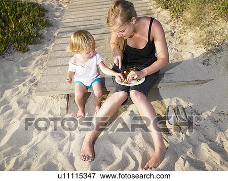 Stock Photo - Woman sitting with a young boy eating at the beach.. Fotosearch