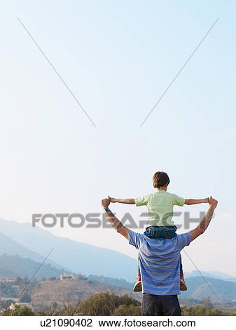 Stock Image - Father carrying son (6-8) on shoulders, rear view. Fotosearch