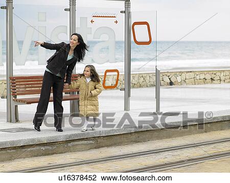 Stock Image - Mother and daughter (6-8) waiting at tram stop on coast. Fotosearch