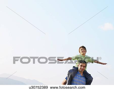 Stock Photo - Son (6-8) sitting on father's shoulders, arms outstretched. Fotosearch