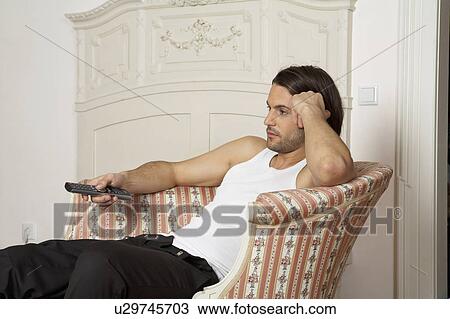 Stock Image - young man sitting on chaise longue holding remote control. Fotosearch