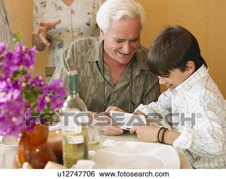 Stock Photograph - Boy (8-10) and grandfather playing portable video game at dining table. Fotosearch