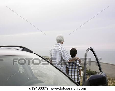 Stock Photo - Boy (8-10) and grandfather standing beside car at beach, rear view. Fotosearch
