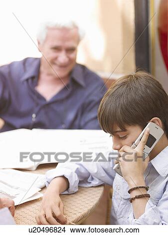 Boy (8-10) sitting at café table, using mobile phone, grandfather looking on View Large Photo Image Picture - Boy (8-10) sitting at café table, using mobile phone, grandfather looking on. Fotosearch
