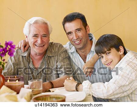 Stock Image - Boy (8-10) sitting at dining table with father and grandfather, smiling, portrait. Fotosearch