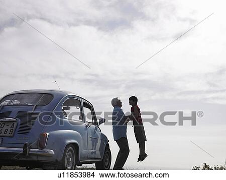 Picture - Senior man lifting grandson (8-10) beside parked car at beach. Fotosearch