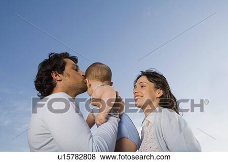 Stock Photo - Couple holding a baby outdoors with the man kissing him.. Fotosearch