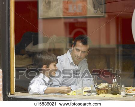 Stock Image - Father and son (8-10) dining in café, view through window, side view. Fotosearch