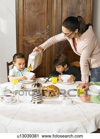 Stock Image - Mother pouring cereal for boy (6-8) and girl (4-6) at breakfast. Fotosearch