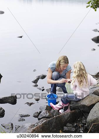 Picture - Woman and young girl collecting pebbles by a lake.. Fotosearch