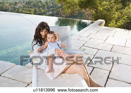 Stock Photograph - Woman holding a baby by an infinity pool smiling.. Fotosearch