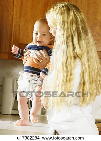 Picture - Woman holding baby up on kitchen counter smiling.. Fotosearch