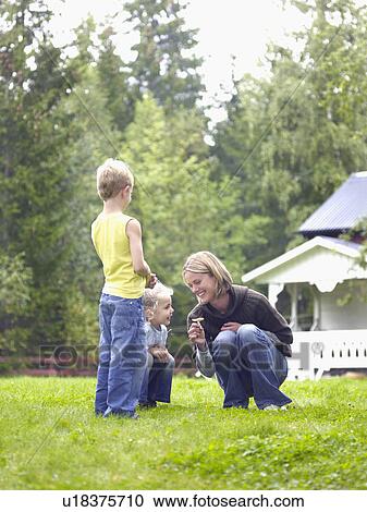 Stock Image - Woman with young girl and young boy in yard picking flower smiling.. Fotosearch