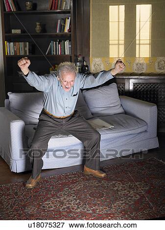 Stock Photo - Grey haired man leaping off sofa in front of television. Alicante, Spain.. Fotosearch