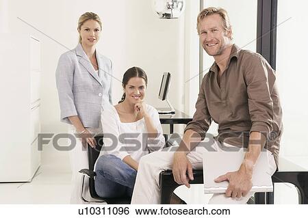 Stock Photograph - Man and two women in office man seated on desk.. Fotosearch