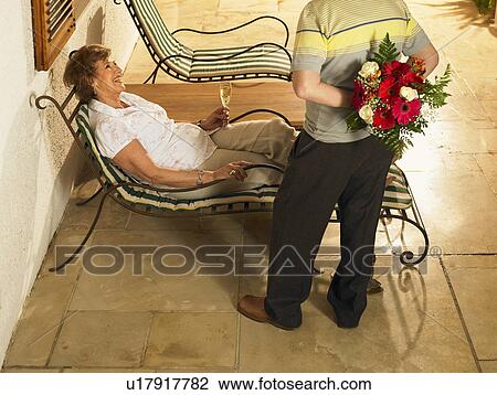 Stock Image - Senior woman lying on sun lounger smiling at senior man with flowers behind back. Fotosearch