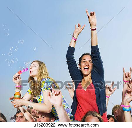 Stock Image - Two girls sitting on shoulders in a crowd blowing bubbles. Fotosearch